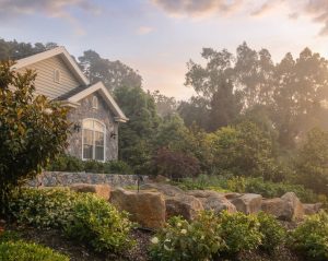 Contemporary home with stone facade and cream siding nestled among large boulders, lush green plantings, and mature forest trees in English cottage garden setting at sunset
